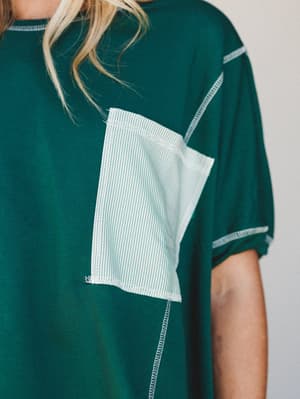 A close-up detail shot of a forest green tee with a contrasting striped chest pocket and exposed overlock stitching — the kind of intentional, lived-in construction detail that makes a basic feel anything but.