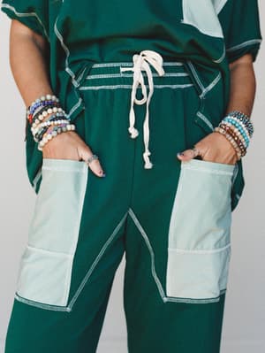 A close-up torso shot of a deep forest green matching set with contrast stripe patchwork pockets and a cream drawstring waist, styled with an armful of layered beaded bracelets that bring the whole thing to life.