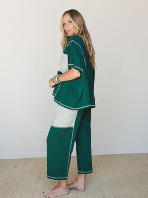 A relaxed two-piece matching set in deep forest green with cream color-block panels and contrast stitching — worn barefoot and styled with stacked beaded bracelets, the whole look reads like a cozy morning that somehow made it onto a mood board.
