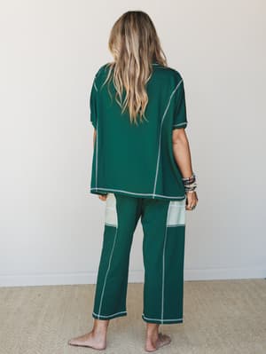 Shot from behind, a model wears a forest green matching pajama set with contrast white topstitching and cream color-block side panels on the pants — relaxed, cozy, and surprisingly put-together.