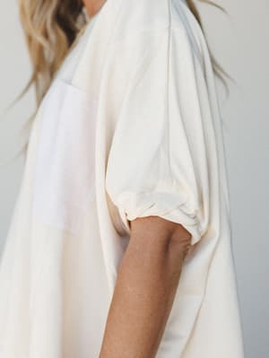 A close-up detail shot of a cream-toned short sleeve top with a gathered, elasticized sleeve hem and a contrasting white patch pocket — soft, relaxed, and quietly thoughtful in its construction.