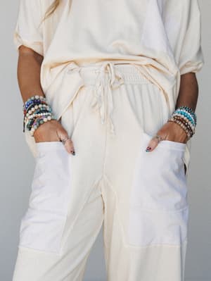 A close-up mid-shot of a cream matching set — relaxed tee tucked loosely into wide-leg drawstring pants with contrast ribbed pocket panels — both wrists stacked with colorful beaded bracelets, hands tucked casually into pockets.