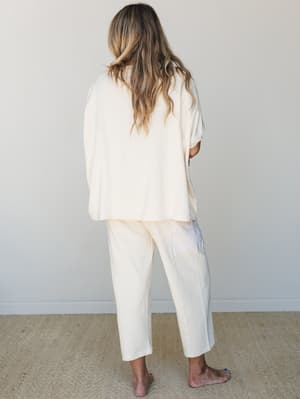 A back-facing shot of a model in a matching cream pajama set — relaxed oversized top and straight-leg pants — photographed barefoot against a soft neutral wall, giving off that slow-morning, nowhere-to-be energy.