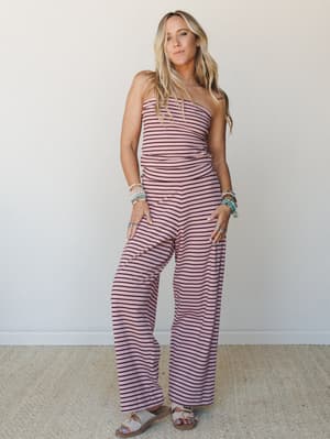 A strapless wide-leg striped jumpsuit in mauve and burgundy worn with layered beaded bracelets and flat sandals — effortless and a little retro, like she's already halfway to somewhere fun.