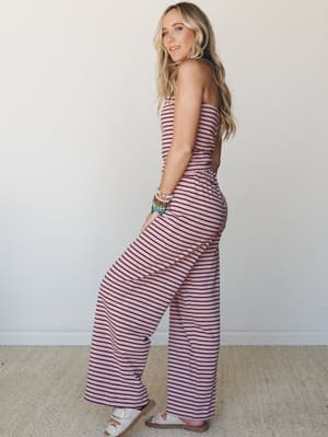 A striped strapless jumpsuit in dusty mauve and deep burgundy with wide-leg trousers, styled with white sandals and a stacked bracelet set — the kind of outfit that looks like you tried just enough.