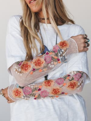 A close-up detail shot of sheer floral embroidered sleeves layered over a white tee — arms crossed to show off the full garden of embroidery, worn with stacked rings and a long necklace that feel very much like hers.