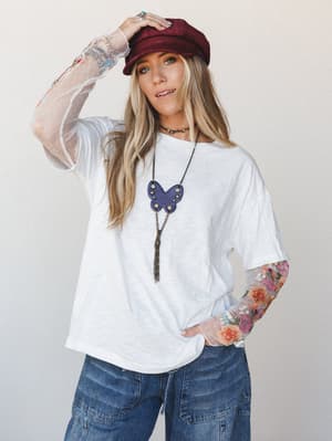 A relaxed off-white tee gets a serious personality upgrade thanks to sheer mesh sleeves covered in colorful floral embroidery — styled with wide-leg denim, a burgundy baker boy cap, and a statement butterfly pendant necklace for a look that's effortl