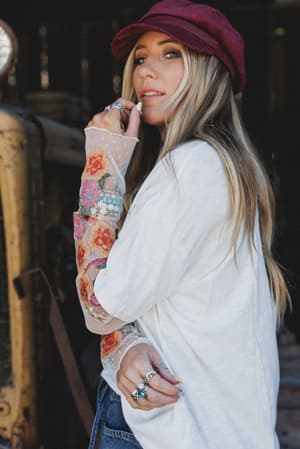 A model looks over her shoulder in a relaxed off-white top with show-stopping embroidered mesh sleeves bursting with floral patches — styled with a burgundy baker boy cap and stacked silver rings against a moody rustic barn backdrop.