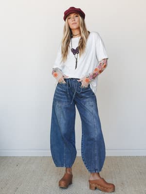 A relaxed, lived-in outfit built around a floral embroidered mesh-sleeve top tucked into voluminous barrel-leg jeans — styled with a burgundy newsboy cap and wooden clogs for that effortlessly artsy, thrift-store-but-make-it-intentional energy.
