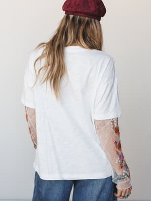 Shot from behind, this layered look pairs a relaxed white tee over sheer, floral-embroidered mesh sleeves — the kind of unexpected detail you only notice when someone walks away, and then you immediately want to know where they got it.