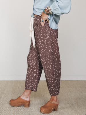 A side-angle mid-shot of barrel-leg printed pants in deep brown with scattered dusty rose spots, styled with a light blue billowy top, wood-soled clogs, and stacked boho jewelry — earthy and effortless.