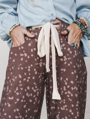 A close-cropped detail shot showing printed barrel jeans in deep brown with a scattered dusty pink floral spot print, styled with a light chambray top and stacked boho jewelry — the cream rope tie at the grommet waist is the kind of detail you notice