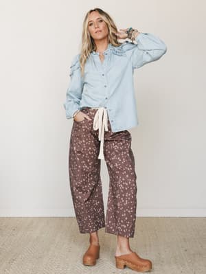 A relaxed full-body shot of a model in a ruffle-trim chambray button-down tied at the hem, paired with wide-leg barrel jeans in a dark brown spotted print, finished with tan wooden clogs and stacked boho bracelets — the kind of outfit that looks effo