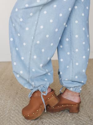A close-up detail shot of light denim polka dot jogger-style pants with tie ankles, styled with tan studded clogs on a woven jute rug — playful, lived-in, and very much the pants you didn't know you needed.