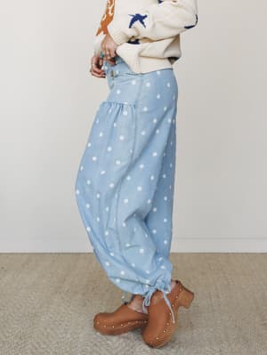 A side-profile shot of relaxed, polka dot denim pants with gathered tiers and tie ankles, styled with a cream intarsia knit sweater and brown studded clogs — the kind of outfit that feels nostalgic and personal all at once.