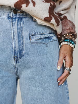 A close-up detail shot capturing light wash barrel jeans styled with a cow print knit sweater tucked at the waist, the look finished with a stacked mix of beaded bracelets, a silver statement ring, and floral nail art — the kind of effortless layerin