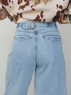 A close-up back view of light wash wide-leg barrel jeans with a vintage-style back buckle closure, styled with a cream and brown cow print sweater and stacked boho bracelets.