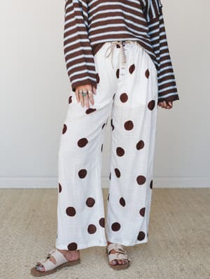 Wide-leg linen pants in off-white with oversized chocolate polka dots, styled with a striped crochet sweater and buckle sandals — the kind of effortlessly playful outfit that looks like it came together on purpose.
