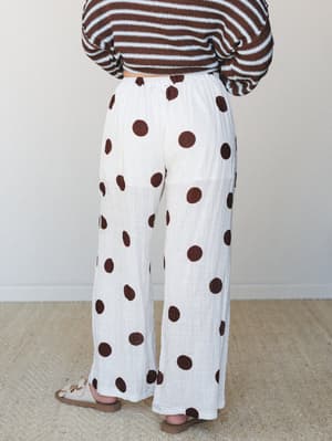 A back-view shot of wide-leg linen-textured pants in off white with oversized terracotta polka dots, styled with a brown and white striped crochet crop sweater and tan strappy sandals — the kind of effortless, print-on-print combo that makes you feel