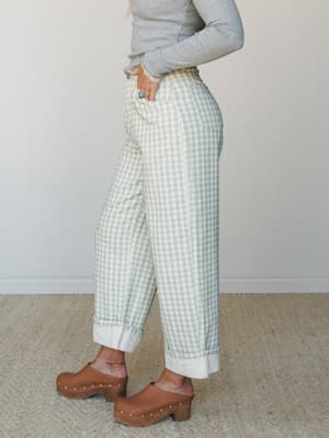 A side-profile shot of wide-leg gingham barrel pants in sage and cream, cuffed at the ankle and paired with a grey long-sleeve and wood-platform clogs — the kind of outfit that makes a Tuesday feel intentional.