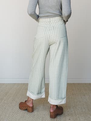 Shot from behind, a model pairs sage gingham wide-leg pants with a fitted grey long-sleeve and brown wooden clogs — relaxed, earthy, and the kind of outfit that looks effortless but was definitely thought about.