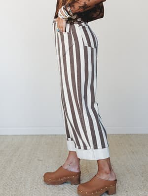 A side-profile shot of wide-leg striped pants in brown and cream, styled with a sheer floral top, stacked beaded bracelets, a turquoise ring, and studded wood clogs — the kind of outfit that looks effortless but was absolutely thought about.