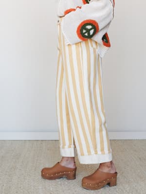 A side-angle crop of wide-leg striped pants with a cuffed hem, styled with a cream novelty knit sweater featuring orange and green peace sign appliqués and paired with brown studded clogs — the kind of outfit that has a whole personality.