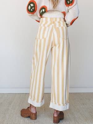 Shot from behind, this model wears wide-leg striped pants in warm golden yellow and cream with rolled cuffs, styled with a cream crochet sweater featuring bold orange and green circle motifs and paired with brown wooden clogs — the whole look reads r
