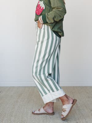 A side-profile shot of wide-leg striped trousers in sage and cream paired with a washed olive graphic sweatshirt and birkenstock-style sandals — effortlessly relaxed with just enough personality.