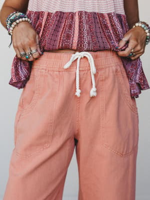 A close-up waist-down shot of relaxed wide-leg pants in a washed peachy-coral tone, styled with a boho printed ruffle top and layered bracelets — the kind of outfit that looks effortless but very much on purpose.