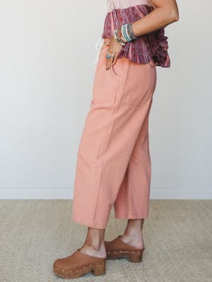 A side-profile shot of wide-leg cropped pants in a warm dusty coral, styled with a paisley ruffle top, stacked bracelets, and wood-heeled clogs — the kind of effortless outfit that looks like it came together in five minutes but definitely didn't.