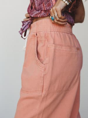A close-up detail shot of wide-leg pants in a warm peach-coral tone, styled with a purple paisley top and stacked boho jewelry — the kind of outfit that feels effortless but absolutely intentional.