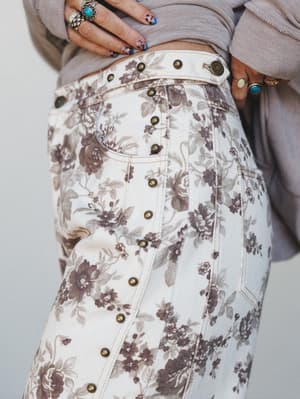 A close-up detail shot of cream floral-print barrel jeans with antique brass stud embellishments running down the side seam, styled with a soft lavender-grey cropped sweater and stacked silver and turquoise rings.