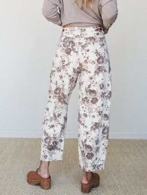 Shot from the back, a model wears wide-leg floral barrel jeans in cream with dusty mauve roses, paired with a tucked taupe long-sleeve top and wooden clogs — the kind of outfit that looks effortless but clearly wasn't an accident.