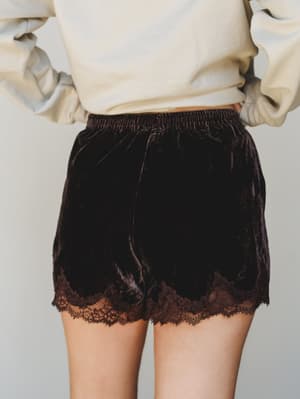 A close-up back view of deep chocolate velvet shorts with a delicate lace hem trim, styled casually with a cream sweatshirt — the kind of piece that blurs the line between cozy-at-home and actually-going-somewhere.