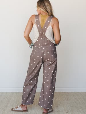 A back-view shot of wide-leg polka dot overalls in mocha brown styled over a cream crochet tank, with layered turquoise bracelets and slide sandals giving the whole look an easy, expressive boho feel.