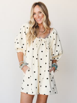 A cream polka dot romper with billowy puff sleeves and a front tie neckline, styled with stacked boho bracelets and layered necklaces — playful and relaxed with a vintage-casual ease.