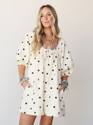 A cream and black polka dot romper with billowy puff sleeves and a tie neckline, styled with stacked turquoise and beaded bracelets for a playful, boho-casual feel against a soft studio backdrop.