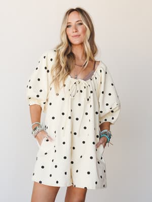 A cream and black polka dot romper with billowy puff sleeves and a tie neckline, styled with stacked turquoise and beaded bracelets for a playful, boho-casual feel against a soft studio backdrop.