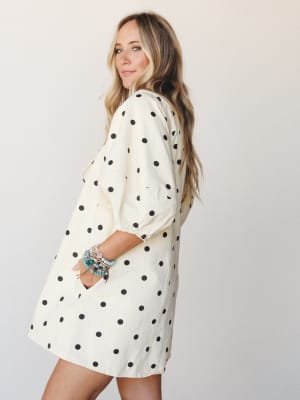 A breezy cream and black polka dot mini romper shot from the back three-quarter angle, styled with a stack of turquoise and beaded bracelets — playful and easy, like something you'd grab off the chair on your way out the door.