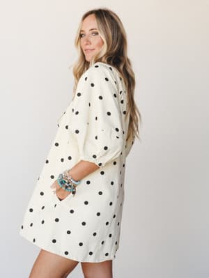 A breezy cream and black polka dot mini romper shot from the back three-quarter angle, styled with a stack of turquoise and beaded bracelets — playful and easy, like something you'd grab off the chair on your way out the door.