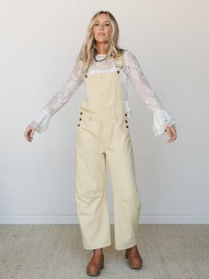 Wide-leg cream overalls styled over a sheer lace long-sleeve top with ruffle cuffs and finished with brown wooden clogs — a free-spirited layering moment that feels vintage-cool without trying too hard.