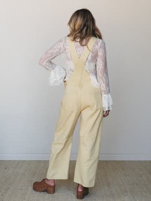Shot from behind, a model wears wide-leg butter-yellow overalls layered over a sheer white lace top with ruffle bell sleeves, finished with brown wooden clogs — a easy, vintage-leaning outfit that feels like a farmers market Saturday done really well