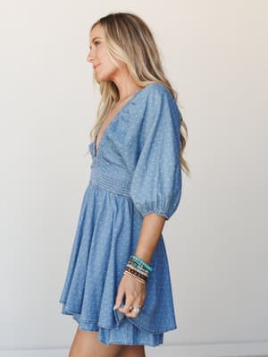 A chambray mini romper in soft denim blue with a white polka dot print, styled with stacked beaded bracelets — it's the kind of piece that looks equally at home at a farmers market or a rooftop happy hour.