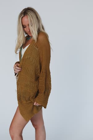 A woman with long blonde hair wears the Brenden Knit Netted Cardigan in brown over a white outfit, standing against a plain light gray background and looking down while holding one side of the cardigan.