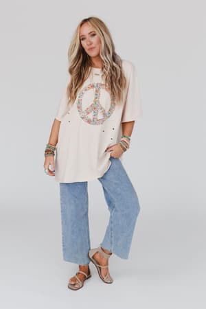 A woman with long blonde hair wears the Jude Oversized Peace Sign Graphic Tee in oatmeal, paired with light blue wide-leg jeans, tan sandals, and colorful bracelets, standing against a plain white background.