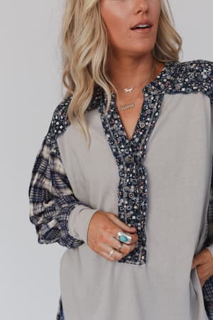 A woman in the Better Together Mixed Print Top - Blue Gray with floral and plaid accents, layered rings including a turquoise one, and a gold “Mama” necklace. She has wavy blonde hair and neutral makeup.