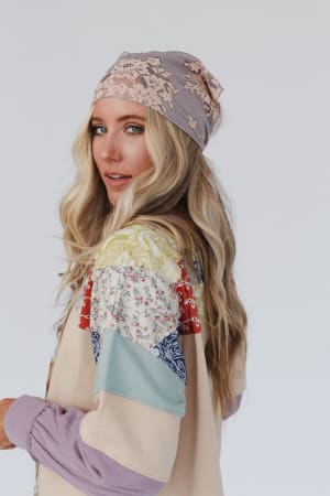 A woman with long, wavy blonde hair wears a pastel patchwork jacket and the Sarah Floral Lace Headscarf in gray, turning over her shoulder against a plain white background.