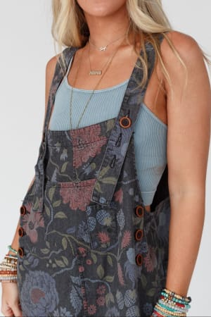 A woman wears High Hopes Slouchy Denim Overalls in Floral Black Denim with a blue ribbed tank top, beaded bracelets, a “MAMA” necklace, and long blond hair; her face is mostly out of frame.