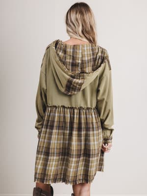 Person wearing a plaid dress with an olive green hooded jacket on a plain background
